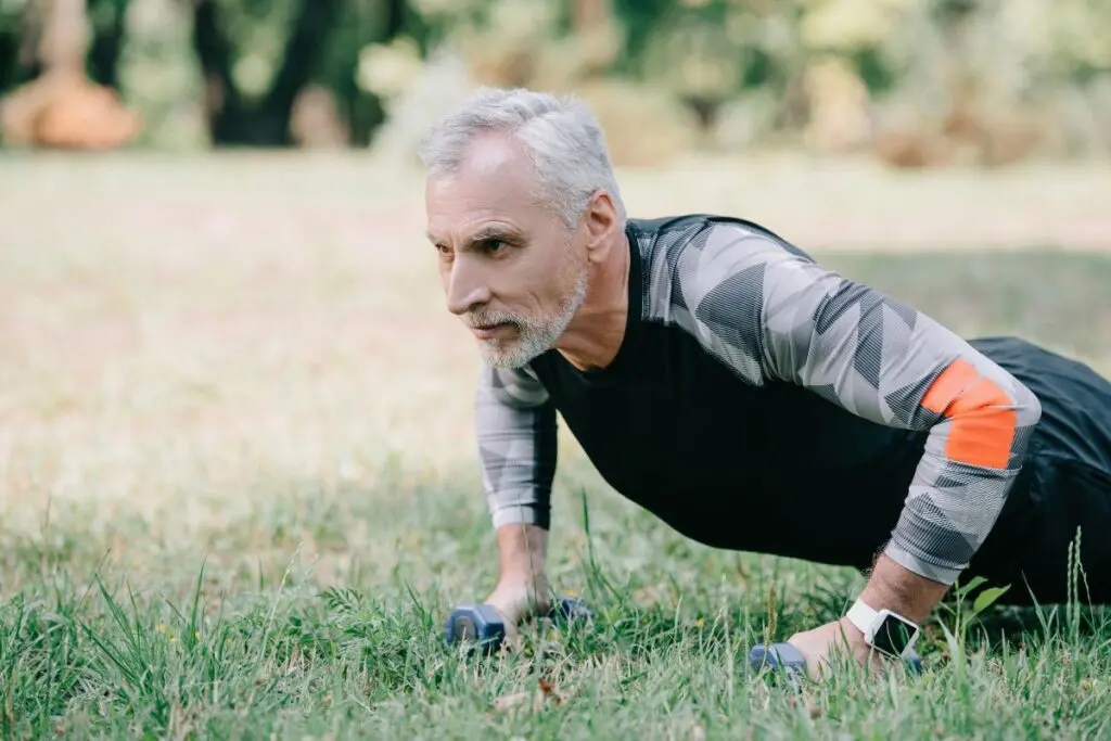 An older man with gray hair and a beard is doing push-ups on a grassy area, wearing a black athletic shirt with gray patterns and an orange sleeve.