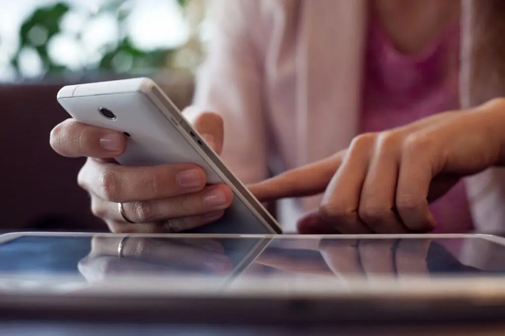 A close-up of a person's hand holding a smartphone while another finger is tapping on the screen of a tablet, with a blurred background of a plant.