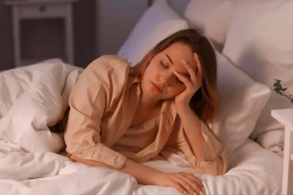 A woman with long hair resting in bed, appearing tired or contemplative, surrounded by white pillows and blankets.