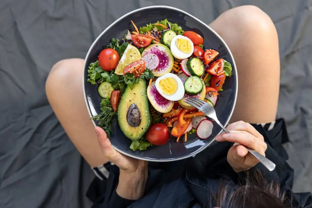 A person holding a plate of fresh salad containing avocado, mixed greens, cherry tomatoes, cucumber, radish, boiled eggs, and sliced vegetables, while sitting on a gray surface.