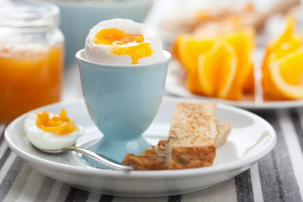 A soft-boiled egg in a light blue egg cup, with a spoonful of egg yolk and a slice of toasted bread on a plate, with orange slices and a jar of orange jam in the background.