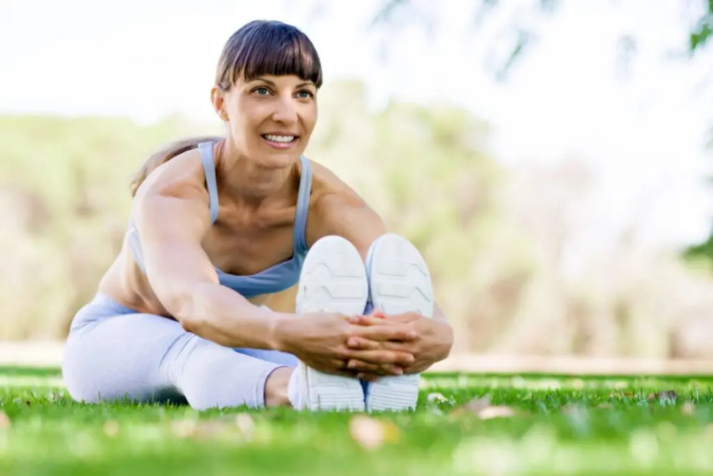 A woman is stretching on a grassy field, holding her feet while smiling.