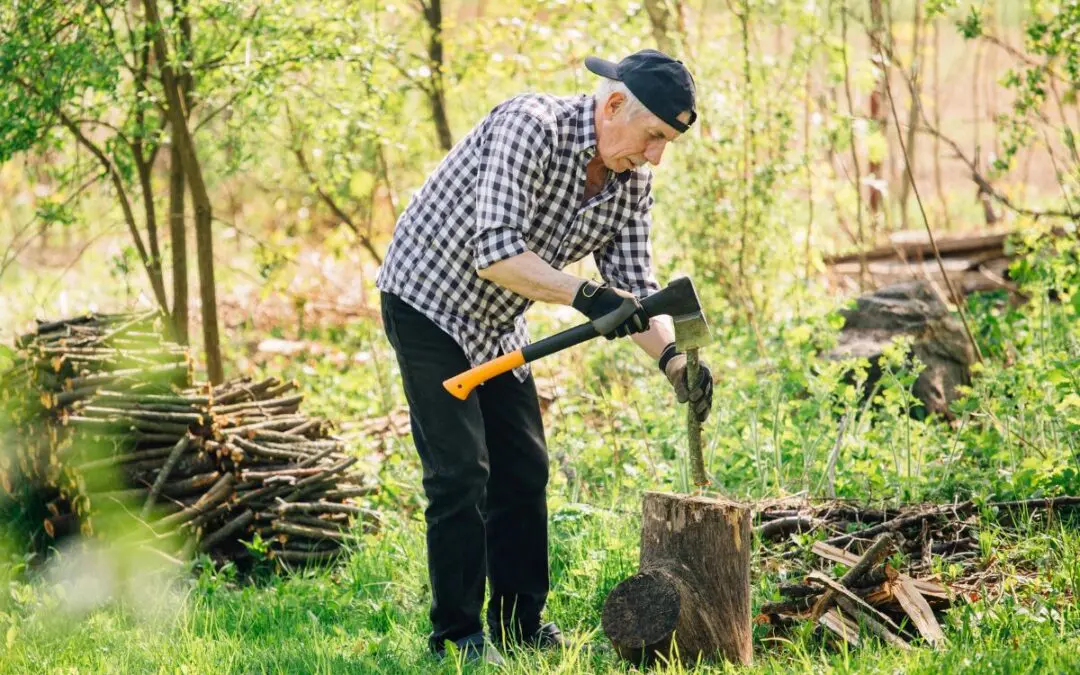 A man chopping wood outdoors with an axe, wearing gloves and a cap, surrounded by trees and stacked firewood.