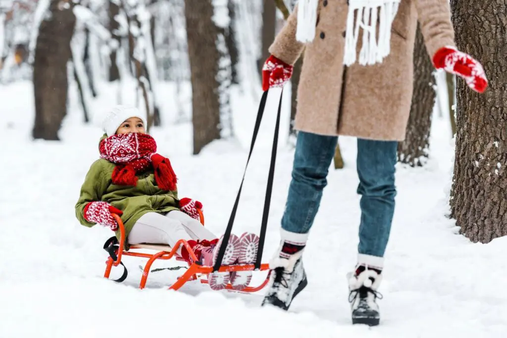 A child sitting in a sled is being pulled through the snow by an adult. The child is bundled up in winter clothing with a scarf and hat, while the adult is wearing a coat, gloves, and boots. Snow-covered trees are visible in the background.