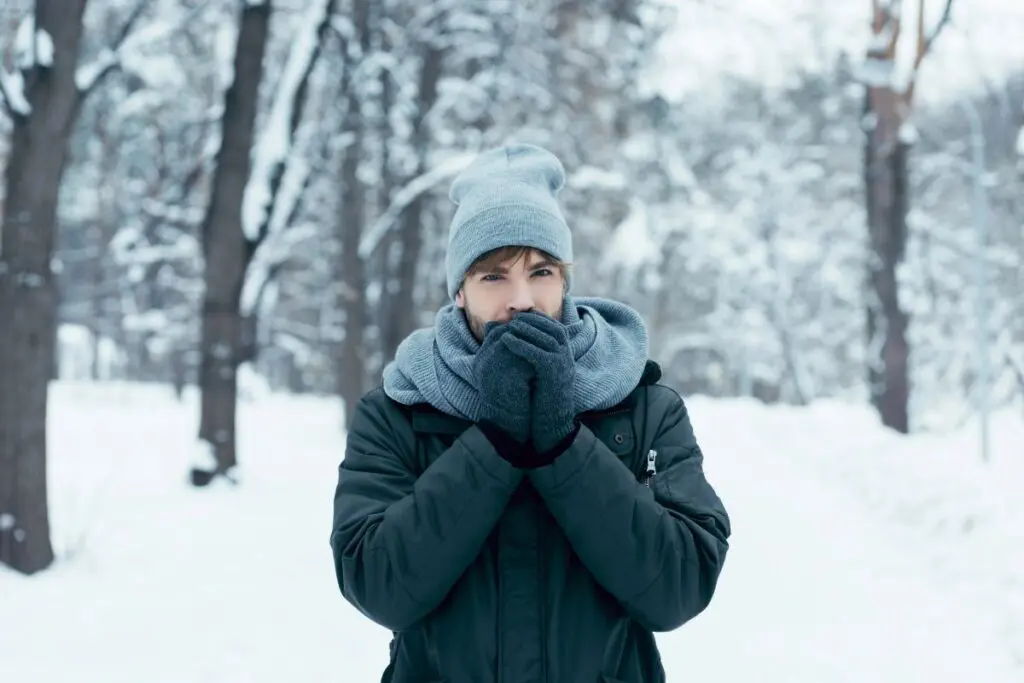A person bundled up in winter clothing, including a gray beanie and scarf, stands in a snowy landscape, holding their hands to their face as if to warm them. Trees covered in snow are visible in the background.