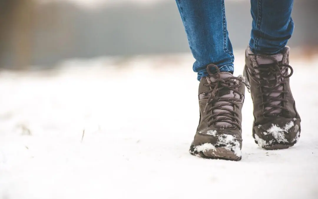 A person walking in the snow, wearing brown hiking boots and blue jeans.