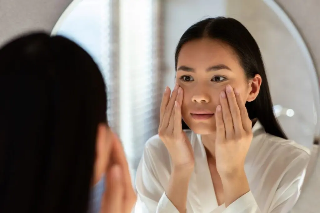 A woman in a white robe applies skincare products to her face while looking in a mirror.
