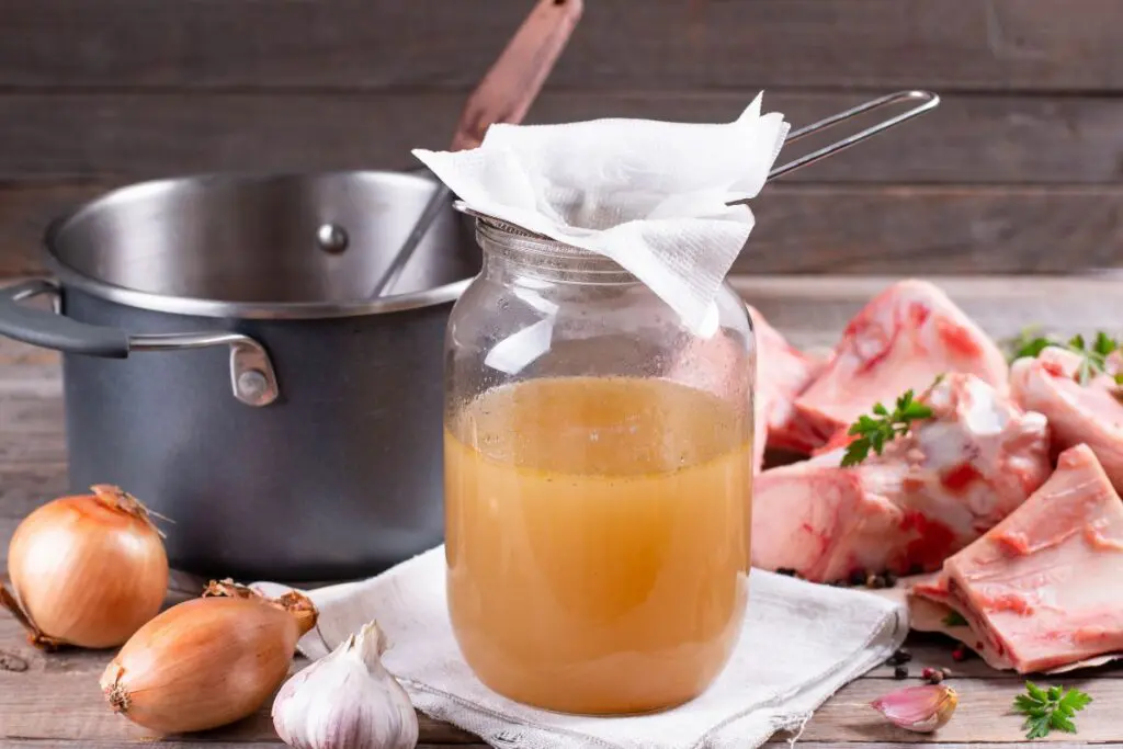 A jar of broth with a strainer on top, surrounded by onions, garlic, a pot, and pieces of meat on a wooden surface.