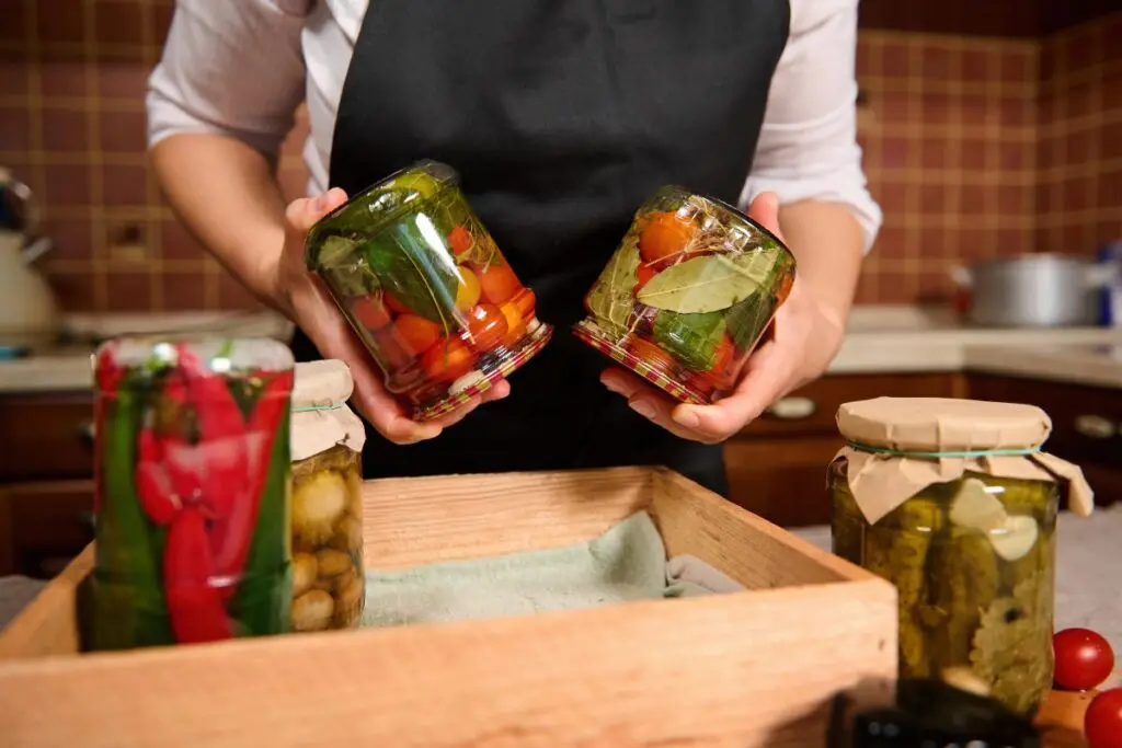 A person holding two jars filled with pickled vegetables, with additional jars and ingredients visible in a wooden crate on the table.