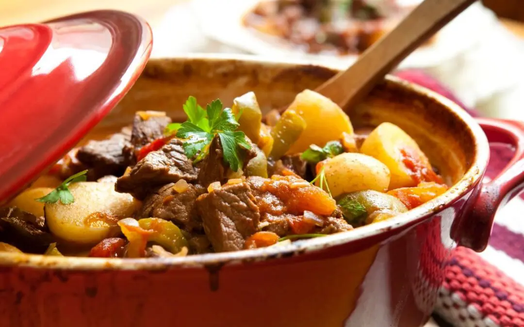 A red pot filled with a hearty beef stew containing chunks of meat, potatoes, vegetables, and herbs.