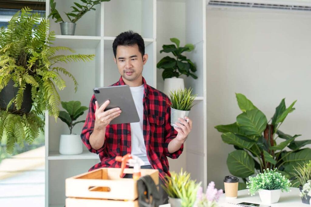 A young man in a flannel shirt examines a potted plant while looking at his tablet.