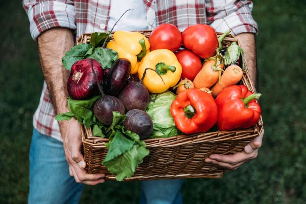 A person holding a wicker basket brimming with fresh vegetables set against a lush, natural background.