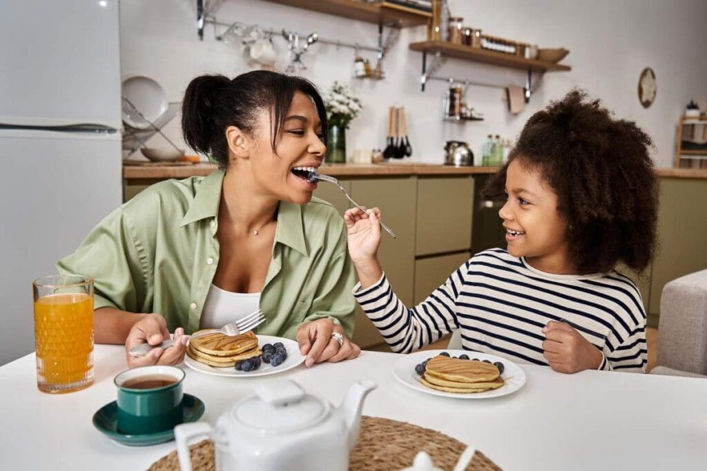 A young girl feeds her mother a blueberry as they sit together at a kitchen table with pancakes, juice, and coffee.
